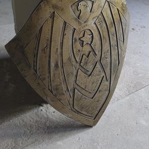 May include: A bronze-colored shield with an eagle design at the top and a star in the center. The shield has a weathered appearance, suggesting it is aged or has been used. The shield is likely a decorative item or prop.