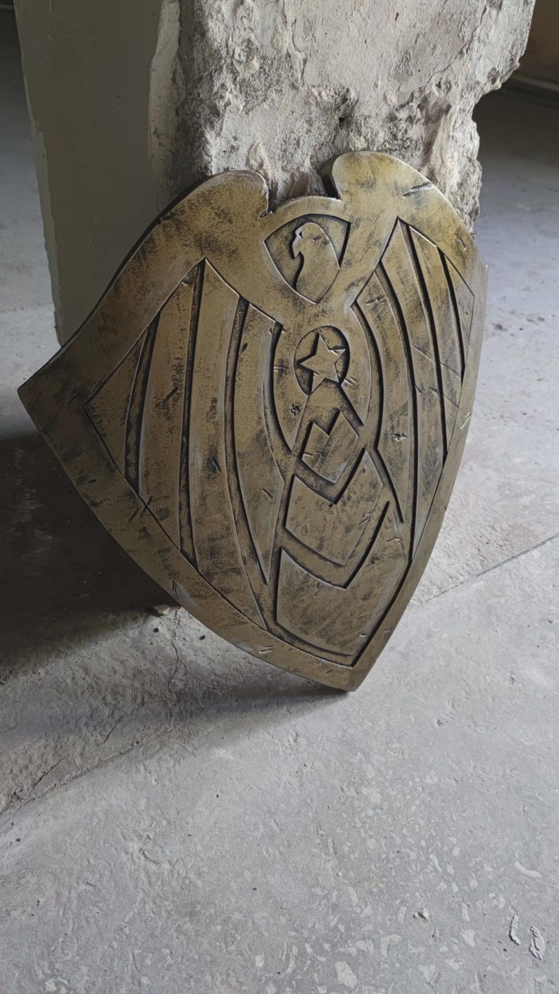 May include: A bronze-colored shield with an eagle design at the top and a star in the center. The shield has a weathered appearance, suggesting it is aged or has been used. The shield is likely a decorative item or prop.