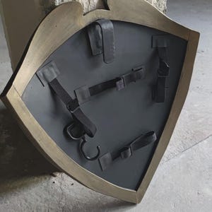 May include: A medieval-style shield with a dark gray surface and a gold-colored trim. The back of the shield features black straps and a hook for carrying. The shield is designed with a unique shape, suggesting a historical or fantasy theme.