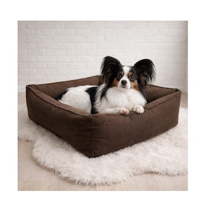Puede incluir: Una cama rectangular marrón para perros con un aspecto afelpado y texturizado. Un perro pequeño con marcas negras, blancas y canela descansa dentro de la cama. La cama está sobre una alfombra blanca y esponjosa.