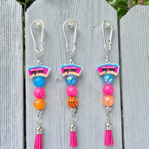 Op de afbeelding: Drie sleutelhangers met een zilveren karabijnhaak, een San Antonio Spurs logo-bedel, kleurrijke kralen in blauw, roze en oranje, en een felroze kwastje. Eén sleutelhanger bevat een kleine basketbal-kraal. De bedels zijn levendig en veelkleurig.