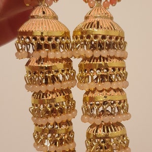 May include: A pair of gold-tone earrings with peach-coloured beads and a layered design. The earrings are made of metal and have a dangling design.