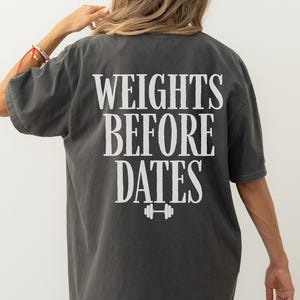 May include: Dark gray t-shirt with the text "WEIGHTS BEFORE DATES" in white block letters. A barbell graphic is below the text. The shirt has short sleeves and a relaxed fit.