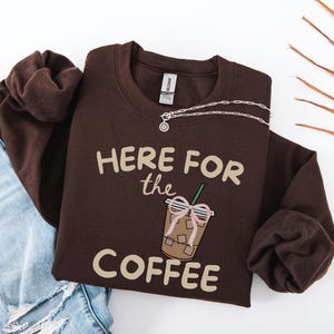 Könnte beinhalten: Ein braunes Sweatshirt mit dem Text "HERE FOR the COFFEE" und einer Grafik eines Eiskaffees mit einer rosa Schleife. Eine silberne Halskette liegt auf dem Sweatshirt, das auf einer blauen Jeans und einem braunen Ärmel liegt.