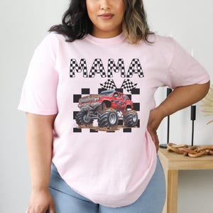 以下が含まれることがあります： 黒と白の市松模様のピンクのTシャツ。赤いモンスタートラックと黒い文字で「MAMA」と書かれている。