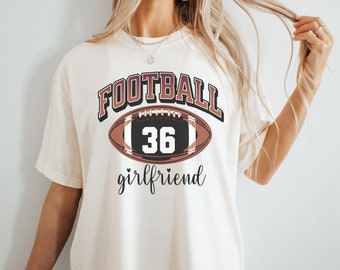 Könnte beinhalten: Cremefarbenes T-Shirt mit einem Football-Motiv. Das Motiv zeigt das Wort "FOOTBALL" in Burgunderrot und Schwarz, eine Footballform mit der Nummer "36" und das Wort "girlfriend" in Schreibschrift.
