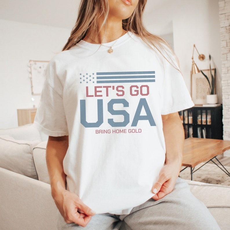 Peut inclure: T-shirt blanc avec un motif bleu et rouge qui dit "Let's Go USA" et "Bring Home Gold".