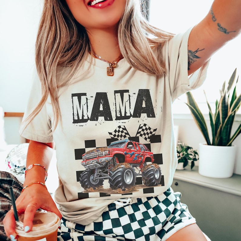 May include: Beige t-shirt with the word "MAMA" in a distressed black font. A red monster truck graphic is below the text, set against a checkered flag background. The shirt has short sleeves.