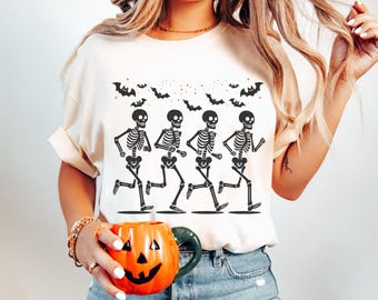 May include: Cream-colored t-shirt featuring a black graphic of four running skeletons and bats. The skeletons are in motion, with their arms and legs extended. The bats are flying above the skeletons. A pumpkin-shaped mug is held in the hand.