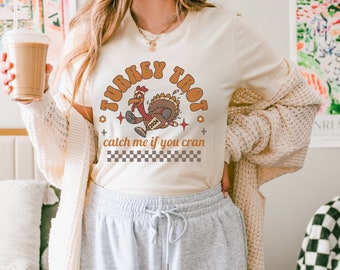 May include: A white t-shirt with a brown turkey running with the text "Turkey Trot 2024" and "catch me if you cran" printed on it.
