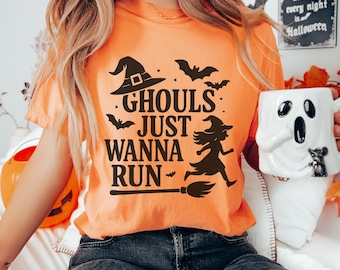 May include: Orange t-shirt with the text "Ghouls Just Wanna Run" in black, featuring a witch on a broom and bat silhouettes. The shirt is worn with dark blue jeans. A white ghost-shaped mug is held in the hand.