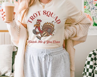 May include: A white t-shirt with a red and brown cartoon turkey running with the text "Trot Squad" and "Catch Me if You Cran" printed on it.