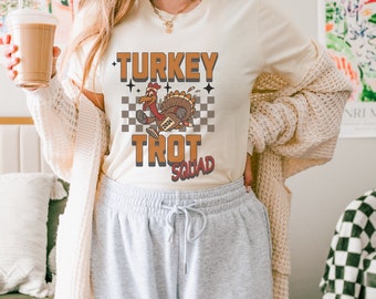 May include: A cream colored t-shirt with a graphic of a cartoon turkey running with the text "TURKEY TROT SQUAD" and the number "2024" on the turkey.