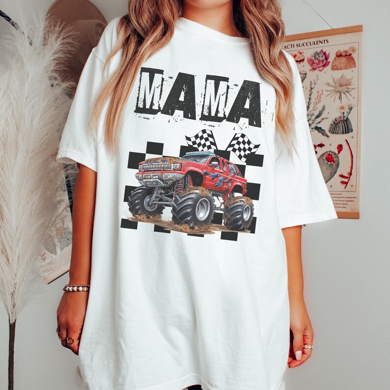 May include: White t-shirt with a distressed "MAMA" graphic above a red monster truck design. The truck is on a checkered background with checkered flags. The shirt is a casual, oversized fit.