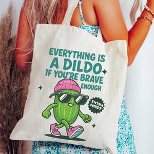 Può includere: Borsa tote color crema con la scritta "EVERYTHING IS A DILDO IF YOU'RE BRAVE ENOUGH" e un cartone animato di cetriolo con occhiali da sole e un berretto rosa. Il cetriolo sta camminando e ha scarpe da ginnastica rosa. La borsa ha anche un adesivo che dice "EXTRA MOIST".