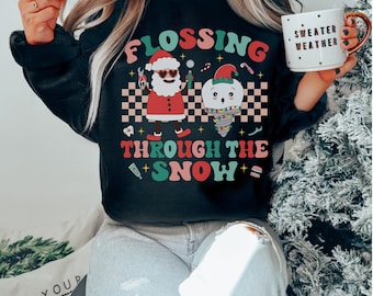 May include: A black sweatshirt with a festive design featuring Santa Claus and a tooth with a Christmas hat. The text reads "Flossing Through The Snow".