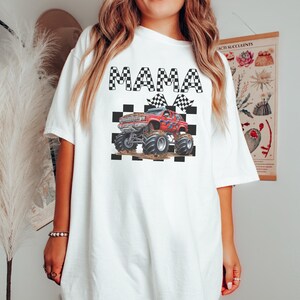 以下が含まれることがあります： 白地に黒と白の市松模様が施されたTシャツ。"MAMA"の文字と、市松模様の旗を持った赤いモンスタートラックが描かれている。