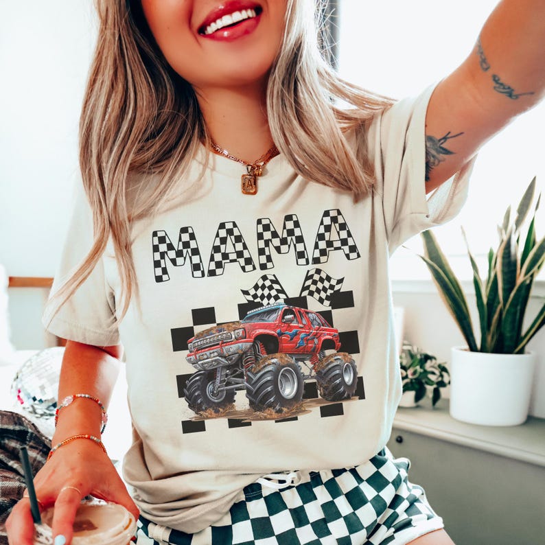 以下が含まれることがあります： 白地に黒と白の市松模様が施されたTシャツ。"MAMA"の文字と、市松模様の旗を持った赤いモンスタートラックが描かれている。