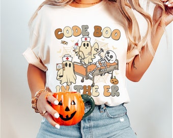 Könnte beinhalten: Ein weißes T-Shirt mit einem Aufdruck einer Geisterkrankenschwester, einem Skelettpatienten und einem Krankenhausbett. Der Text auf dem Shirt lautet "Code Boo in the ER".