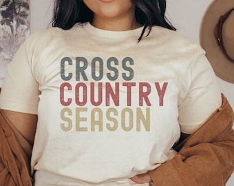 Może przedstawiać: Biały T-shirt z napisem "CROSS COUNTRY SEASON" w stylu vintage. Tekst jest w trzech kolorach: szarym, czerwonym i beżowym.