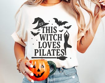 以下が含まれることがあります： 「THIS WITCH LOVES PILATES」の黒いグラフィックがプリントされたクリーム色のTシャツ。魔女の帽子、コウモリ、ヨガのポーズが描かれています。緑色の持ち手が付いたジャック・オー・ランタンのマグカップが手に持たれています。ハロウィーンをテーマにしたアパレルです。