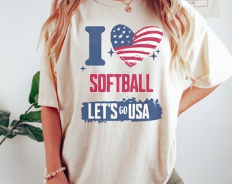 May include: A cream-colored t-shirt with a blue "I" and a red, white, and blue heart with a softball inside. The text "Softball" is in red and "Let's Go USA" is in blue.