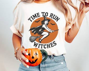 May include: Cream-colored t-shirt featuring a Halloween design. The graphic includes a witch riding a broom inside an orange circle, with the text "TIME TO RUN WITCHES" in a circular arrangement. A small orange pumpkin mug is held in the hand.