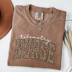 May include: A brown t-shirt with the words "telemetry NURSE" printed on the front. The word "telemetry" is in a cursive font, and "NURSE" is in a leopard print design. A silver necklace with a small pendant is on the shirt.