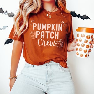 Pode incluir: Uma mulher vestindo uma camiseta cor ferrugem com o texto "Pumpkin Patch Crew" e um gráfico de abóbora. Ela está segurando uma caneca de viagem branca com um padrão de abóbora.