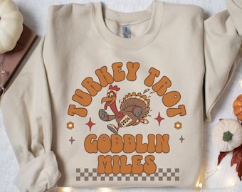 May include: A beige sweatshirt with a retro-style graphic of a running turkey with the text "Turkey Trot Gobblin Miles" and the year "2024" on the turkey's bib.