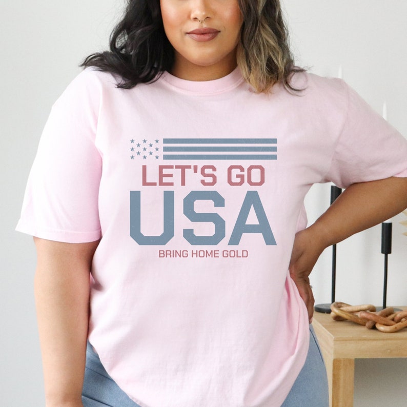 Peut inclure: T-shirt rose avec un motif de drapeau am&eacute;ricain bleu et blanc et le texte "Let's Go USA Bring Home Gold".
