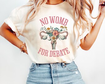 Peut inclure: T-shirt crème avec l'inscription rose "NO WOMB FOR DEBATE" au-dessus d'une illustration florale de l'utérus. Le motif comprend diverses fleurs et feuilles. Le t-shirt est porté avec un jean bleu.