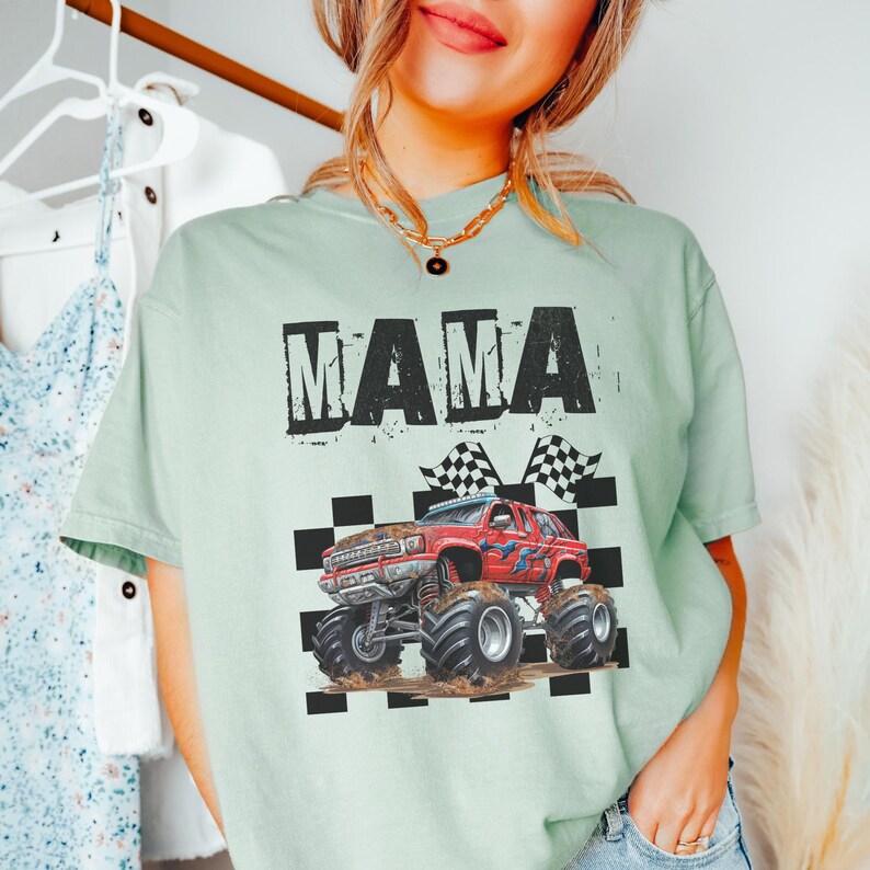 May include: A light green t-shirt with the word "MAMA" in black block letters. Below the text is a red monster truck graphic with checkered flags. The truck is covered in mud.