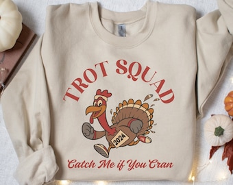 May include: A beige sweatshirt with a cartoon turkey running with the text "Trot Squad" and "Catch Me if You Cran" printed on it. The turkey is wearing a red Santa hat and has a number "2024" on its side.