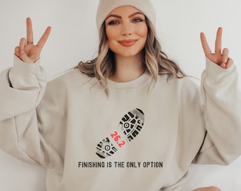 Könnte beinhalten: Ein beiger Pullover mit einem schwarz-weißen Aufdruck eines Laufschuhs mit der Zahl 26,2 in Rot. Der Text "FINISHING IS THE ONLY OPTION" ist unter dem Aufdruck gedruckt.