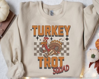 May include: A beige sweatshirt featuring a cartoon turkey running on a chequered background. The text "TURKEY TROT SQUAD" is printed in orange and brown lettering. The turkey has the number "2024" on its side.
