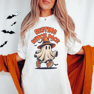 May include: White t-shirt with the text "Resting Witch Face" and a cartoon ghost wearing a witch hat and orange sneakers. Black bat decorations are in the background. The shirt is part of a Halloween-themed collection.