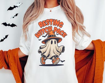 May include: White t-shirt with the text "Resting Witch Face" and a cartoon ghost wearing a witch hat and orange sneakers. Black bat decorations are in the background. The shirt is part of a Halloween-themed collection.