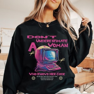 Peut inclure: Sweat-shirt noir avec l'inscription "DON'T UNDERESTIMATE A WOMAN WHO KNOWS HER CODE". Le graphisme représente un ordinateur rétro avec des accents roses et jaunes. Le texte supplémentaire comprend "I'M THE GIRL THEY SAID COULDN'T CODE, BREAKING STEREOTYPES, HACK THE GENDER GAP."