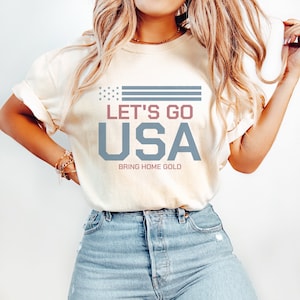 Peut inclure: Un t-shirt blanc avec un motif bleu et rouge qui dit "Let's Go USA" et "Bring Home Gold".
