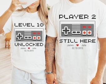May include: Two white t-shirts with a pixelated video game controller design. One shirt says "Level 10 Unlocked" with names and a date. The other shirt says "Player 2 Still Here" with names and a date.