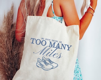 May include: Cream-colored tote bag with the text "NO SUCH THING AS TOO MANY Miles" in blue. Below the text is a blue illustration of running shoes. The bag has two cream-colored handles.