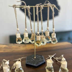 Könnte beinhalten: Ein Set mit 12 klaren Glas-Lufterfrischern für das Auto mit geometrischer Form, die an einem goldenen Metallständer mit schwarzem Marmorsockel hängen. Die Lufterfrischer sind mit einer hellgelben Flüssigkeit gefüllt und haben einen Holzdeckel mit einem geflochtenen Seil.