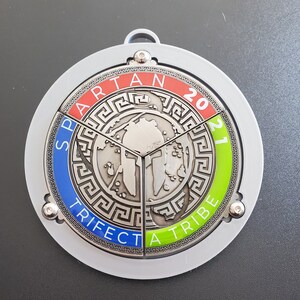 以下が含まれることがあります： スパルタンレースの銀メダル。スパルタンのヘルメットのデザインと、縁に「Spartan Trifecta 2021」の文字が刻まれています。