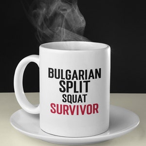 May include: White ceramic coffee mug with black text that reads "Bulgarian Split Squat Survivor" in all caps. The word "Survivor" is in red. The mug is sitting on a white saucer.