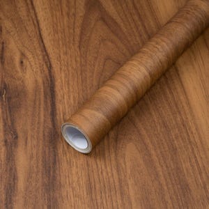 May include: A roll of adhesive contact paper featuring a realistic medium brown wood grain pattern with dark brown streaks and knots. The roll is positioned diagonally across a flat surface that displays the same matching wood grain design.