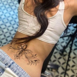 May include: A black tattoo design on the lower back featuring an ornate, symmetrical pattern. The design includes swirling lines, floral elements, and a central jewel-like shape. The person is wearing a white tank top and blue jeans.