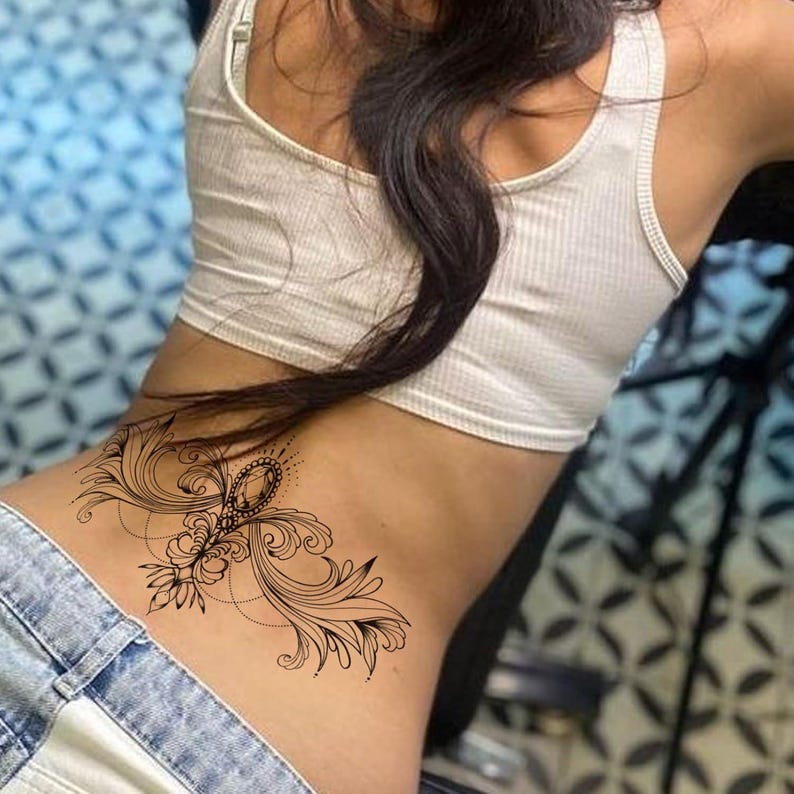 May include: A black tattoo design on the lower back featuring an ornate, symmetrical pattern. The design includes swirling lines, floral elements, and a central jewel-like shape. The person is wearing a white tank top and blue jeans.