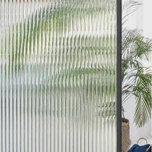 May include: A room divider screen featuring vertical fluted glass panels held within a thin black frame. The textured glass creates a distorted, semi-transparent view of green foliage behind it, while the structure stands upright on a floor.