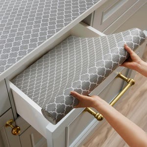 May include: A roll of gray shelf liner featuring a white quatrefoil pattern. The liner is partially unrolled inside a white kitchen drawer with gold-toned hardware. The same gray and white patterned material covers the countertop surface above.